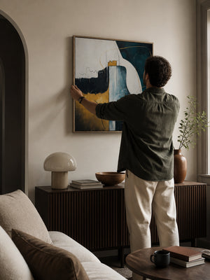 Man hanging a abstract painting on a wall in a living room.